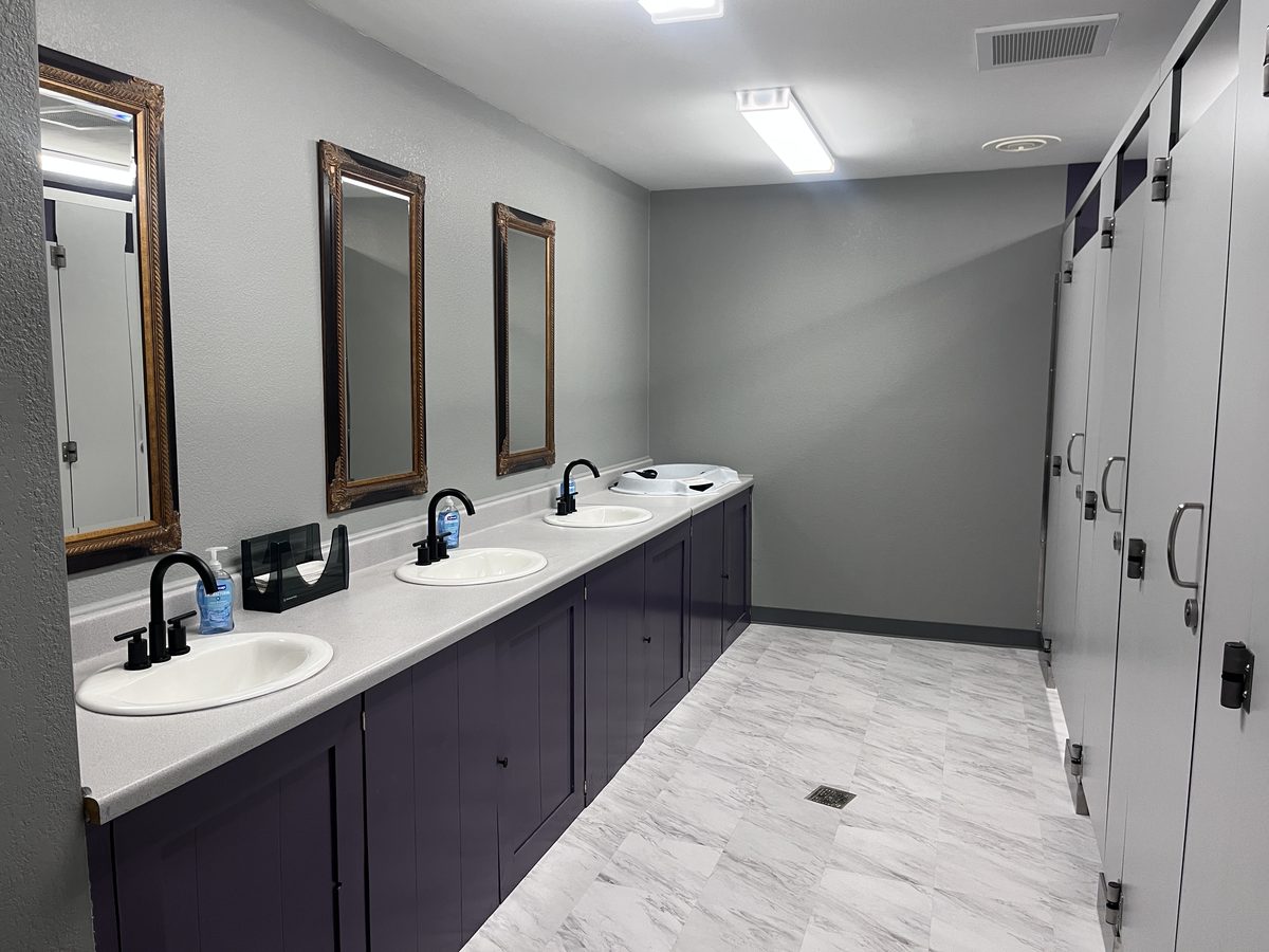 Finished commercial restroom with gray walls, a navy vanity, and gold-framed mirrors